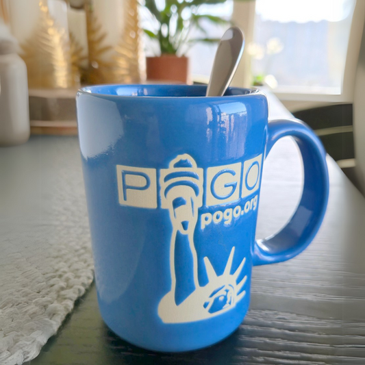 Blue glazed ceramic mug on a table with a spoon inside. The mug has a tall handle and a matte off-white design of unglazed clay showing the Statue of Liberty, POGO logo, and text: pogo.org.