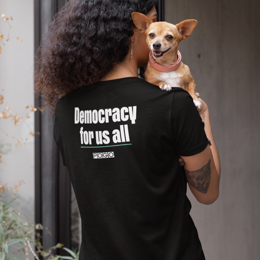 Woman wearing a black t-shirt. On the back of the shirt, centered white text: Democracy for us all and white POGO logo.