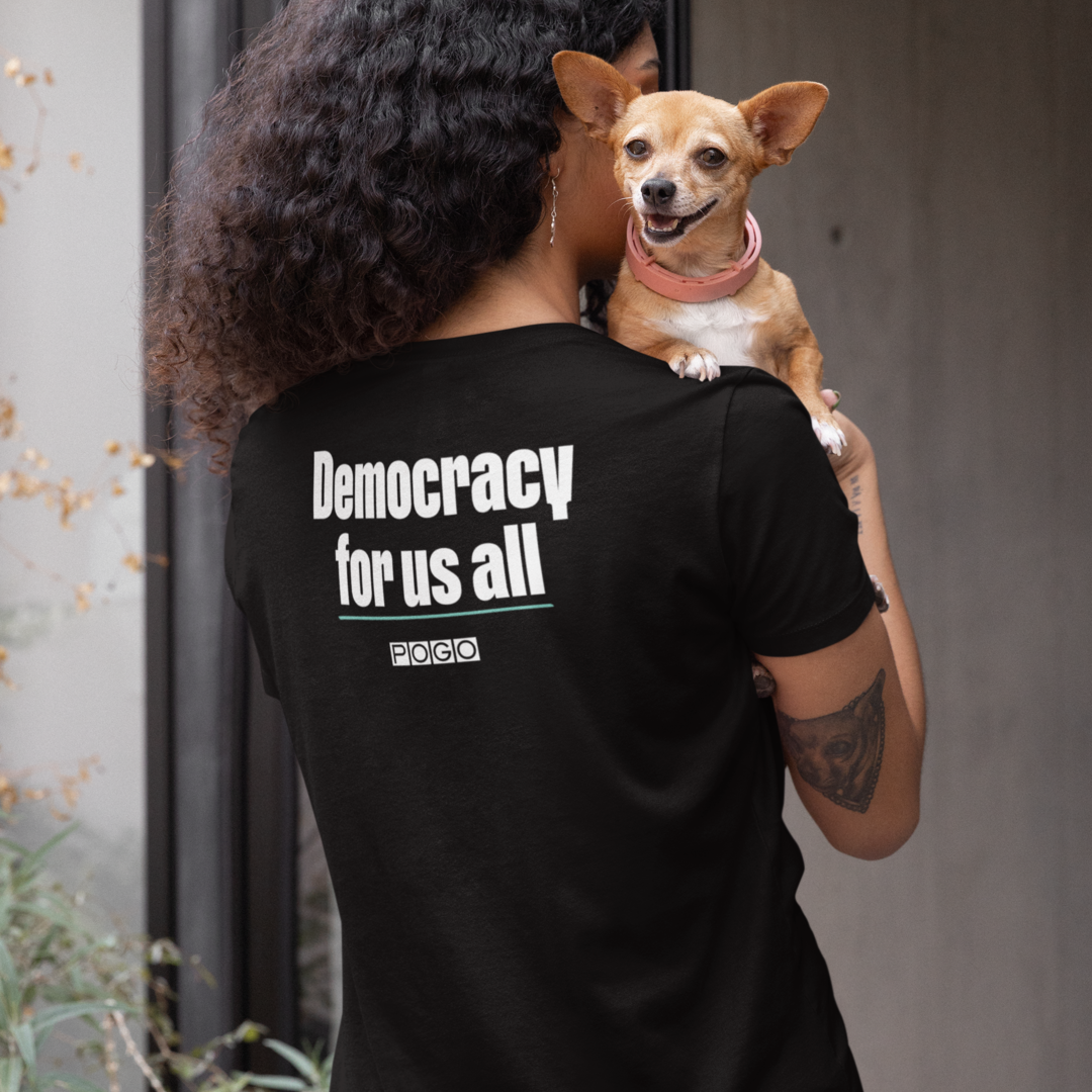 Woman wearing a black t-shirt. On the back of the shirt, centered white text: Democracy for us all and white POGO logo. 